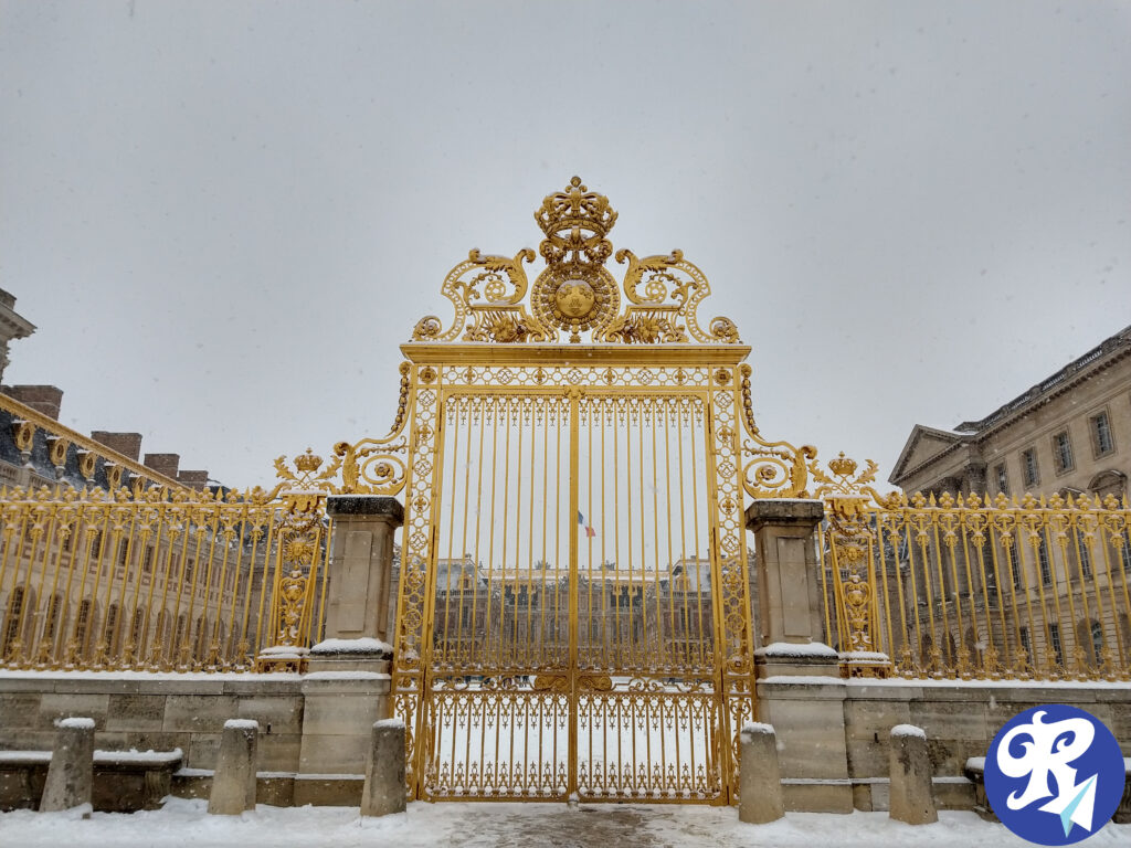 Os portões dourados ornamentados do Castelo de Versailles. Os portões são altamente decorativos, com designs e motivos intrincados, incluindo um emblema central no topo. A cena está ambientada no inverno, como evidenciado pela neve no chão e nos portões. Os edifícios do palácio são visíveis ao fundo, exibindo uma arquitetura clássica. A imagem também inclui um logotipo no canto inferior direito, um "R" estilizado com um avião de papel, demonstrando o logo do Blog. Esta imagem é interessante e relevante, pois retrata um famoso marco histórico conhecido por sua arquitetura opulenta e sua importância na história francesa.