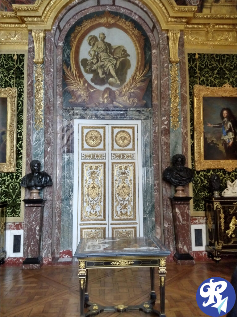 Porta em um dos Cômodos do Castelo de Versailles. Uma sala opulenta com uma porta ricamente decorada no centro. A porta é adornada com desenhos intrincados em ouro e branco, emoldurada por colunas de mármore e um arco. Acima da porta, há uma grande pintura que representa uma cena clássica com uma figura em uma túnica esvoaçante. A sala possui papel de parede ornamentado em verde e dourado, e há dois bustos em pedestais flanqueando a porta, ao lado direito é possível ver um quadro de Louis XIV. Em frente à porta, há uma mesa com superfície escura e detalhes dourados. O piso é de madeira polida com um padrão geométrico.