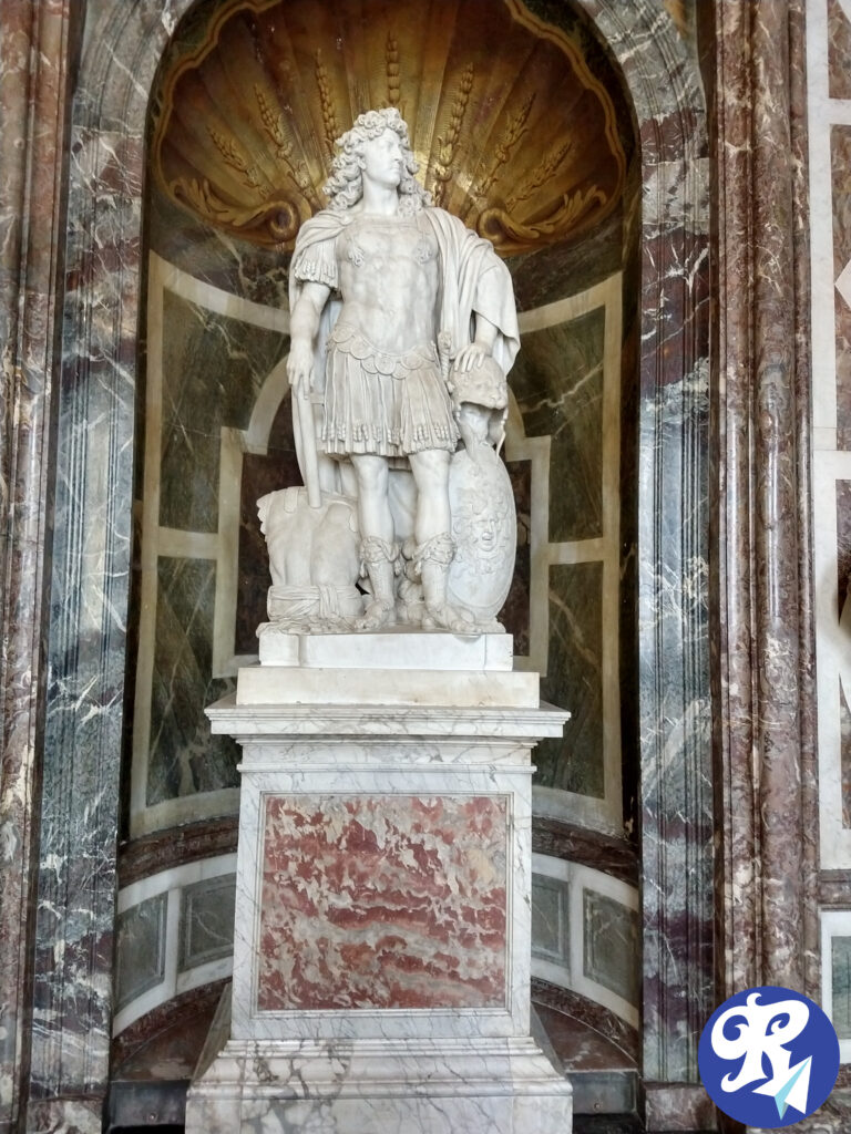 Estátua de Louis XIV, Rei que governou a França de 1.643 até sua morte em 1.715. A imagem mostra uma escultura em mármore de Louis XIV vestido com armadura ao estilo romano, de pé sobre um pedestal. A estátua está localizada em um nicho ornamentado com um design em forma de concha acima dela e é cercada por paredes de mármore com padrões intrincados. A figura é representada com uma mão descansando sobre um escudo e a outra sobre a cabeça de um leão. O pedestal também é de mármore e apresenta um padrão veinado em vermelho e branco.