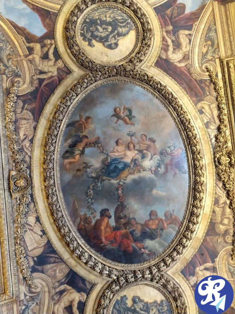 Teto de um dos cômodos de Versailles. Imagem mostra um teto ornamentado com detalhes dourados intrincados e vários painéis pintados. A pintura oval central representa uma cena mitológica com várias figuras, incluindo querubins e deidades, rodeadas por nuvens e guirlandas de flores. As áreas circundantes apresentam elementos decorativos adicionais e cenas menores, todas emolduradas por molduras douradas elaboradas. Esta imagem é interessante e relevante pois mostra a grandiosidade e o detalhamento artístico típico do design interior clássico ou barroco.