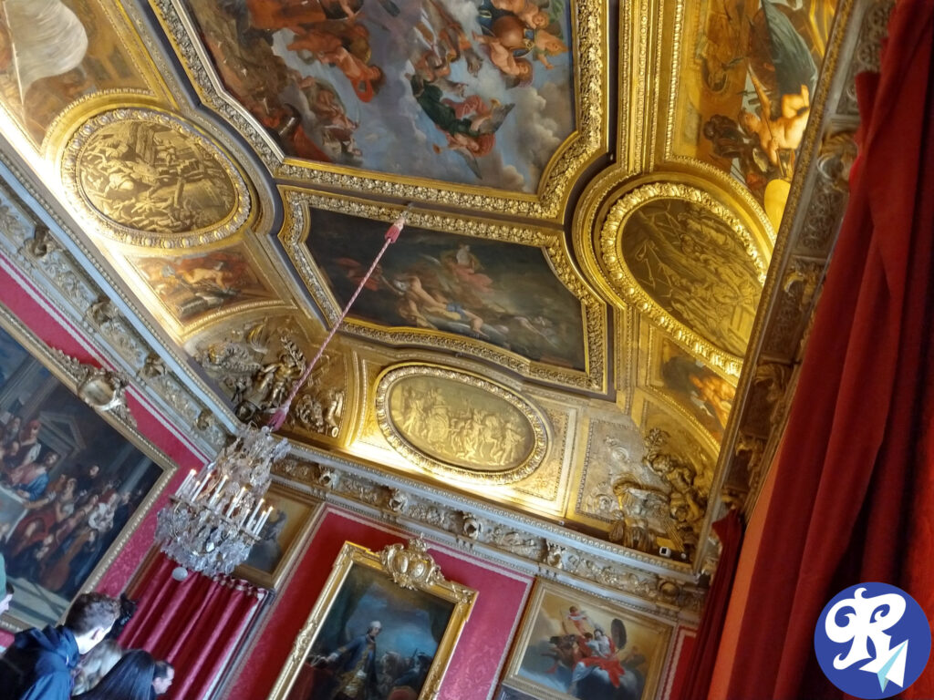 A imagem mostra uma sala opulenta com um teto e paredes ricamente decorados. O teto apresenta detalhes intrincados em ouro e várias grandes pinturas que retratam diversas cenas, provavelmente mitológicas ou históricas. As paredes são adornadas com grandes pinturas emolduradas e cortinas vermelhas. Há um lustre pendurado no teto, adicionando um toque luxuoso ao ambiente. Algumas pessoas são visíveis na parte inferior da imagem. Um dos Cômodos de Versailles