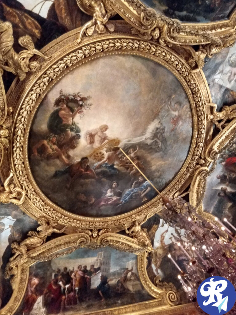 Teto de um dos Cômodos do Castelo de Versailles A imagem mostra um teto ornamentado com decorações douradas elaboradas e pinturas intrincadas. A pintura central retrata uma cena mitológica com várias figuras, incluindo uma pessoa segurando uma coroa de flores e outra figura em uma carruagem puxada por cavalos. Ao redor da pintura central, há pinturas adicionais emolduradas, cada uma representando diferentes cenas, possivelmente históricas ou mitológicas. O teto é ricamente decorado com folhas de ouro e entalhes detalhados, e um lustre é parcialmente visível pendurado no teto. A impressão geral é de opulência e maestria artística