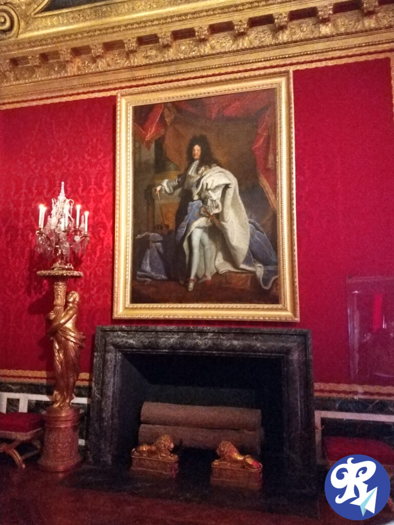 A imagem mostra o famoso retrato de Louis XIV, pintado por Hyacinthe Rigaud em 1701. Louis XIV, também conhecido como o Rei Sol, é retratado em trajes de coroação opulentos. Ele está usando um manto de veludo azul adornado com flores-de-lis douradas, símbolo da realeza francesa. Sob o manto, ele veste uma túnica branca decorada com rendas elaboradas. Louis XIV segura o cetro real em uma mão, enquanto a outra repousa graciosamente sobre a mesa ao lado dele. Ele também está usando peruca e sapatos de salto alto, ambos na moda da época. A pintura é rica em detalhes, destacando o poder e a majestade do rei, e é considerada uma das imagens mais icônicas da monarquia francesa. Abaixo da pintura descrita acima, está uma lareira. À esquerda da lareira, há uma estátua dourada segurando um candelabro com velas acesas. A lareira em si é preta com estátuas de leões dourados na base, e há toras colocadas dentro. O teto também possui decorações douradas elaboradas.