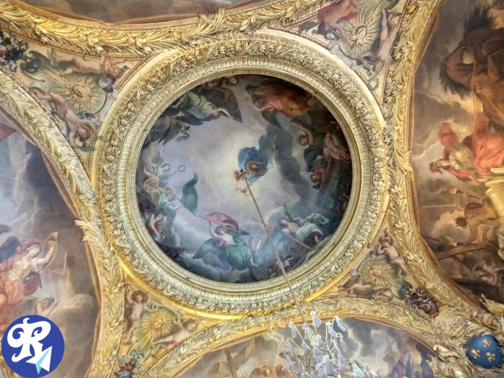 Teto de um dos Cômodos do Castelo de Versailles: A imagem mostra um teto ornamentado com intrincados detalhes dourados e várias pinturas. O teto possui um grande ornamento circular central cercado por desenhos detalhados em folhas de ouro e várias cenas pintadas. As pinturas retratam diversas figuras, incluindo querubins e outras personagens mitológicas ou históricas.