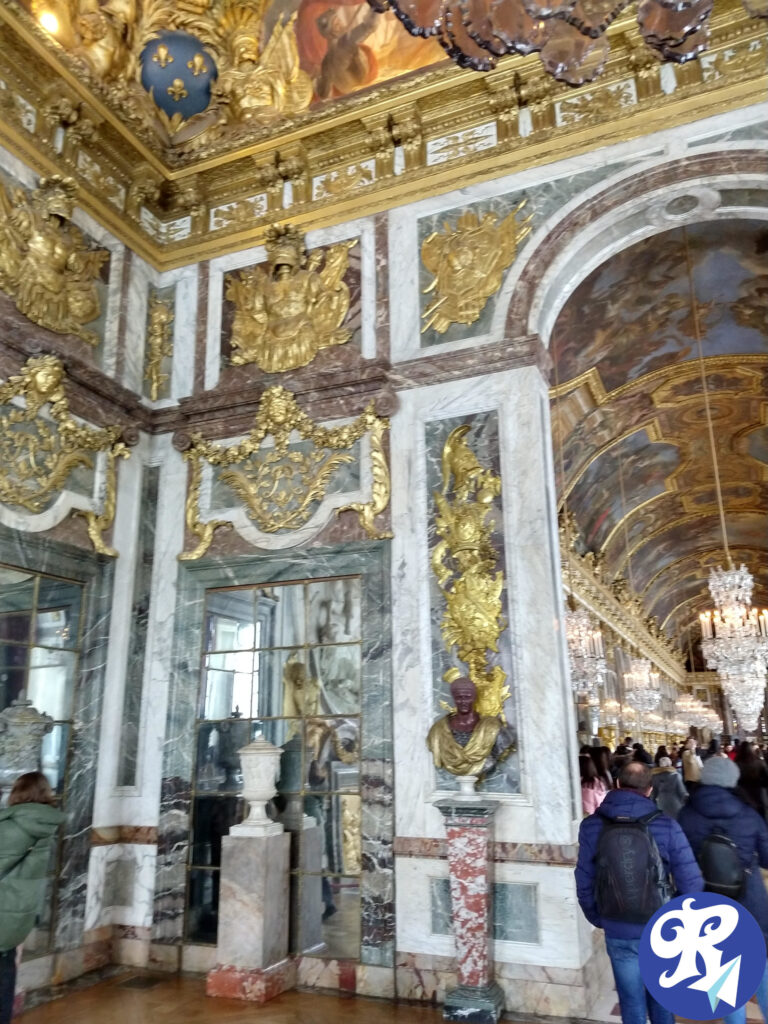 A imagem mostra o interior opulento do Castelo de Versailles, a entrada para a Galeria dos Espelhos. As paredes e o teto são ricamente decorados com intricados adornos dourados, mármore e afrescos detalhados. Existem grandes espelhos, bustos clássicos e urnas ornamentadas colocadas em pedestais. O teto apresenta pinturas elaboradas e decorações em folha de ouro. O salão está cheio de pessoas. O estilo arquitetônico e as decorações indicam uma influência barroca, com um alto nível de artesanato e luxo.