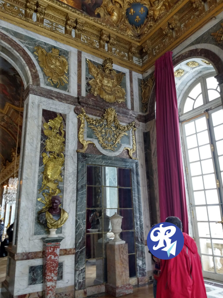 A imagem mostra um interior ornamentado do Caastelo de Versailles, o lado direito da entrada da Galeria dos Espelhos. As paredes são decoradas com entalhes dourados intricados e painéis de mármore. Há várias esculturas em relevo douradas, incluindo uma de uma águia com asas abertas e outra de um escudo com espadas cruzadas. O teto também é ricamente decorado com ouro e arte pintada. Um grande espelho com uma moldura de mármore está embutido na parede, e em frente a ele há um pedestal com uma urna decorativa. A sala possui janelas altas com cortinas vermelhas, permitindo a entrada de luz natural. A impressão geral é de opulência e grandeza, típica de residências reais ou nobres.