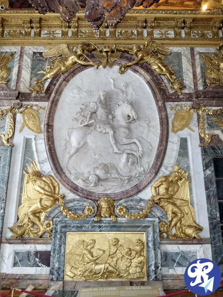 A imagem mostra uma decoração de parede ornamentada com uma grande escultura em relevo oval central, representando Louis XIV a cavalo. Em torno do relevo central, há decorações intrincadas em ouro, incluindo duas figuras aladas no topo e duas figuras sentadas em cada lado, cada uma segurando diferentes objetos. Abaixo do relevo central, há um painel retangular dourado com uma cena detalhada de figuras, incluindo um anjo e várias crianças. A parede é de mármore com acentos dourados e entalhes intrincados. Na parte inferior da imagem, há uma placa com texto em francês e inglês. O texto diz: Salão da Guerra