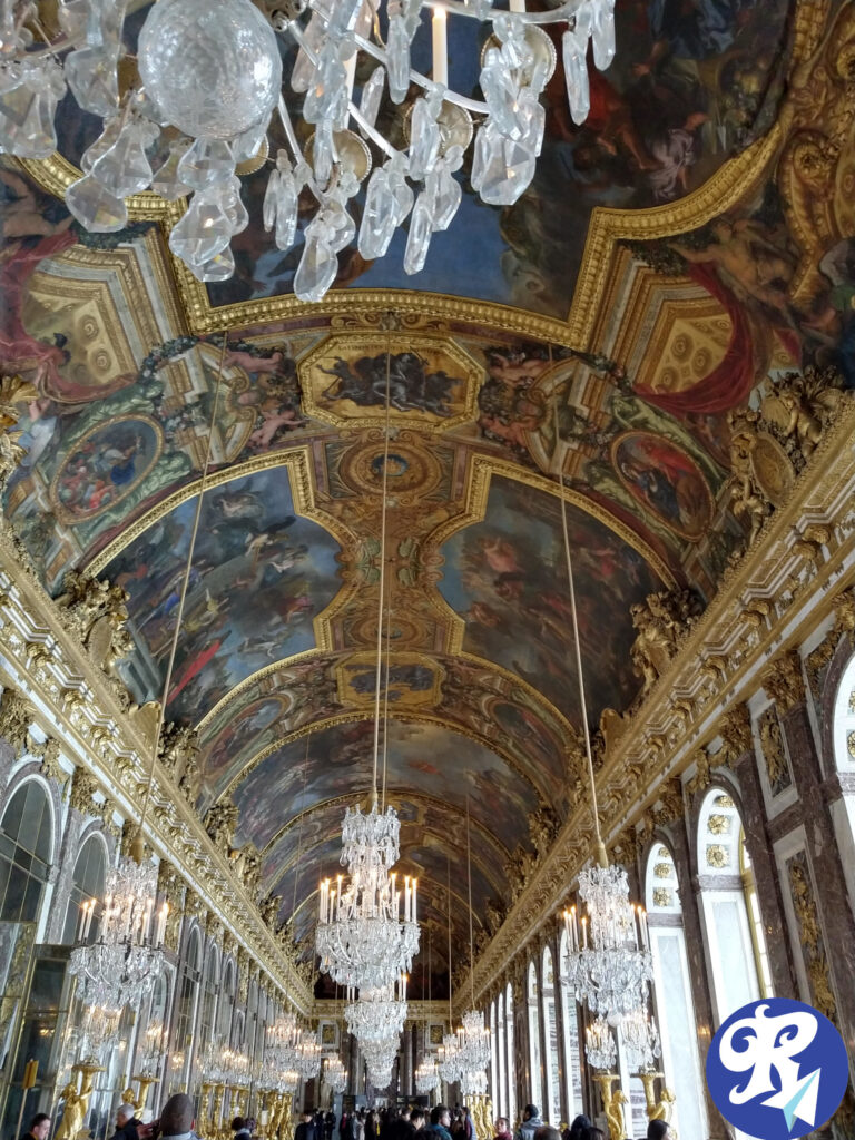 Galeria ou Salão dos Espelhos (Galerie des Glaces, em francês) no Castelo de Versailles. Este salão grandioso é conhecido por seu design opulento, com uma série de grandes espelhos de um lado e janelas do outro, com lustres de cristal pendurados no teto. O teto em si é ricamente decorado com pinturas intrincadas e detalhes dourados. O salão é um marco histórico e arquitetônico significativo, famoso por seu papel em diversos eventos históricos, incluindo a assinatura do Tratado de Versalhes em 1919. A imagem captura a natureza luxuosa e ornamentada do salão, destacando sua importância artística e histórica.