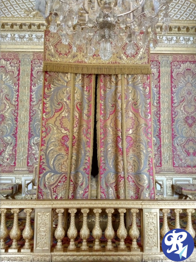 O Quarto do Rei A imagem mostra o Quarto do Rei no Castelo de Versailles com uma decoração opulenta. No centro, há uma cortina ornamentada que envolve a cama do Rei com padrões florais em tons de dourado, vermelho e azul, pendurada em uma estrutura dourada. Acima da cortina, há um lustre de cristal com várias peças penduradas, refletindo a luz. Abaixo da cortina, há um parapeito dourado com detalhes esculpidos. As paredes ao redor também são ricamente decoradas com padrões dourados e vermelhos, complementando a cortina central. A imagem transmite uma sensação de riqueza e grandiosidade.