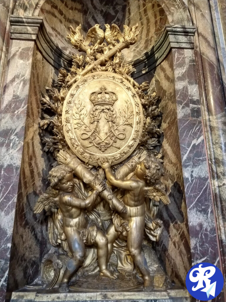 A imagem mostra uma escultura ornamentada e dourada situada dentro de uma alcova de mármore. O elemento central da escultura é um grande escudo redondo adornado com uma coroa e com o monograma de Louis XIV, incluindo cetros cruzados e ramos de louro. Acima do escudo, há duas águias voltadas uma para a outra, segurando um feixe de varas com um machado, conhecido como fasces, simbolizando autoridade e poder. Flanqueando o escudo, há dois querubins ou putti alados, que parecem estar apoiando ou apresentando o escudo. O fundo e a área circundante são ricamente decorados com mármore, adicionando à grandiosidade da cena. Esta imagem é interessante e relevante, pois retrata uma peça de arte altamente detalhada e simbólica