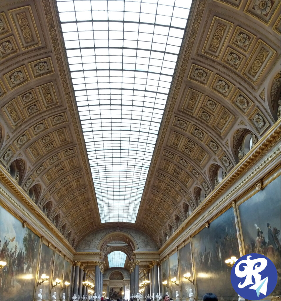 A imagem mostra a Galeria das Batalhas, localizada no Castelo de Versailles. Esta galeria é um longo e majestoso corredor com um teto abobadado e envidraçado, decorado com detalhes dourados e relevos intricados. As paredes são adornadas com grandes pinturas que retratam cenas de batalhas históricas importantes para a França. Essas pinturas são ricamente detalhadas e capturam momentos dramáticos e heróicos da história militar francesa. Além das pinturas, ao longo do corredor há várias esculturas de bustos de figuras históricas, que contribuem para o ambiente solene e imponente da galeria. A iluminação natural entra através do teto envidraçado, complementada por lustres e arandelas nas paredes, destacando as obras de arte e os detalhes arquitetônicos. A decoração e a arquitetura são grandiosas, refletindo a opulência e a importância histórica do Castelo de Versailles. Essa galeria é uma das muitas atrações impressionantes do palácio, sendo um testemunho da rica herança cultural e histórica da França.