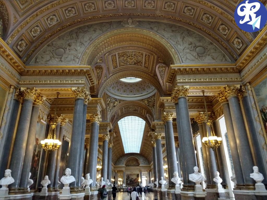 A imagem mostra a entrada da famosa Galeria das Batalhas no Castelo de Versailles. Este majestoso corredor é ricamente decorado com detalhes dourados e mármore. À direita, há grandes colunas de mármore que sustentam a estrutura, e entre elas, podem ser vistos bustos em pedestais, representando figuras históricas importantes. As paredes são adornadas com grandes pinturas que retratam cenas de batalhas significativas na história francesa. O teto, abobadado e ornado com detalhes intrincados, permite a entrada de luz natural através de uma claraboia, destacando as obras de arte e a arquitetura impressionante. A iluminação é complementada por lustres pendurados ao longo do corredor. A grandiosidade e a opulência da galeria refletem o esplendor e a importância histórica do Castelo de Versailles.